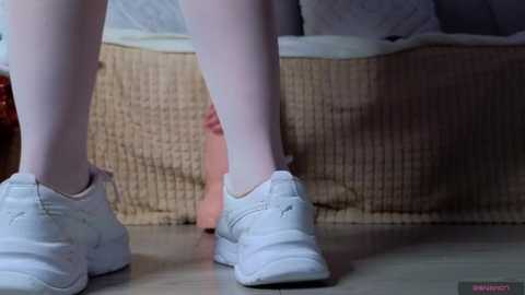 Media: A video of a person\u2019s legs in white socks and sneakers, standing on a wooden floor near a beige wicker basket and a bed with white linens.