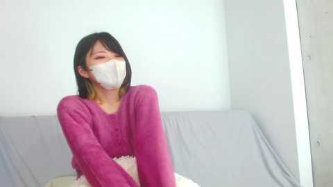 Media: Video of an Asian woman with shoulder-length black hair, wearing a pink fleece hoodie and white surgical mask, sitting on a light grey couch in a brightly lit room.