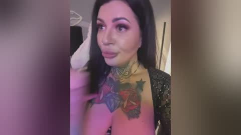 Video of a woman with long dark hair, light skin, and tattoos on her chest and neck. She's wearing a black lace top and has a confident, sultry expression.