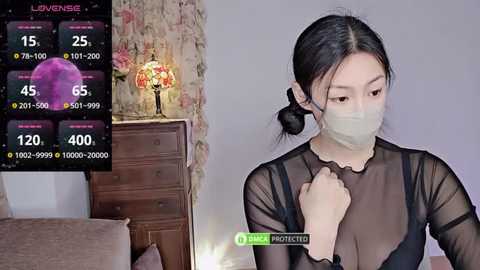 Media: Video of a young Asian woman with light skin and long black hair in a bun, wearing a black sheer top, a face mask, and a watch. Background includes a wooden dresser with a lamp and a floral-patterned wall.