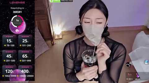 Media: A video of an Asian woman in a transparent black top and face mask, holding a glass of water. Background shows a wooden floor, a bed, and a wall.