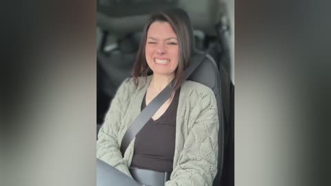 Media: A video of a smiling Caucasian woman with straight brown hair, wearing a black top and a beige cardigan, seated in a car, with a gray seatbelt across her lap.