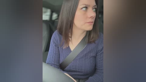 Video of a young woman with shoulder-length brown hair, wearing a blue sweater, seated in a car, looking contemplative.