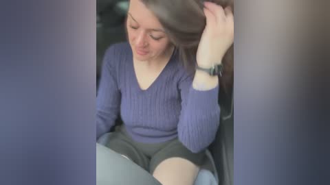 A video of a young woman with long brown hair, wearing a purple ribbed sweater and black shorts, sitting in a car, looking down, with a black wristwatch on her left wrist.