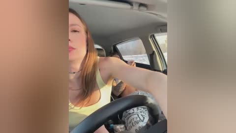 Media: Video of a woman with long brown hair, wearing a white tank top, driving a car with a tattooed arm visible, steering wheel in hand, in daylight.