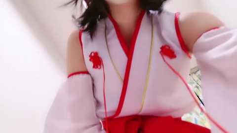 A video of a person in a white and red kimono, with intricate red embroidery, and long, flowing sleeves, standing against a blurred, white background.