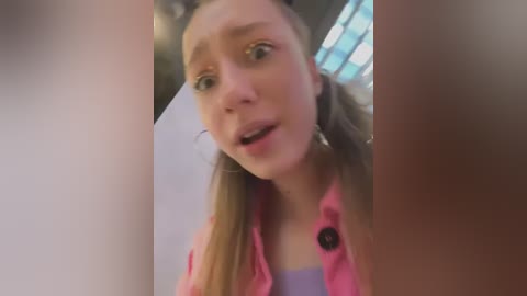 Media: A video of a young woman with fair skin and light brown hair styled in pigtails, wearing a pink jacket and purple top, appearing surprised with wide eyes and open mouth, blurred background.