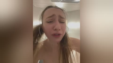 Media: Video of a young woman with light skin, wet hair in pigtails, wearing large hoop earrings, and a distressed expression, taken indoors with a blurry background.