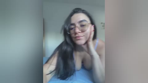 Media: A video of a young woman with long, dark hair, wearing glasses, light blue tank top, and resting her chin on her hand, in a dimly lit room with beige walls.