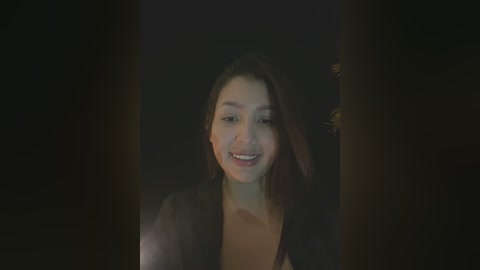 Media: A video of a young woman with fair skin and long, dark hair, smiling with dimples, wearing a black top, set against a dark, blurred background.