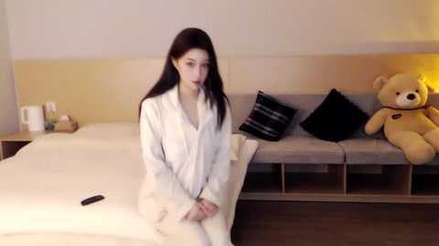 Media: Video of a woman in a white robe sitting on a bed with a beige headboard, surrounded by black and beige pillows, and a teddy bear.