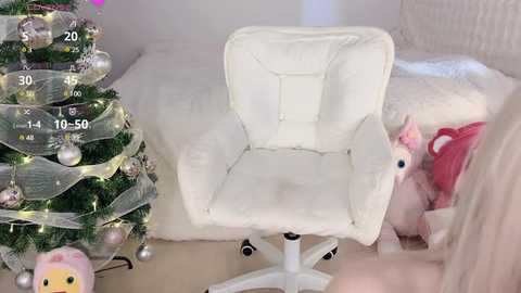 Media: Video of a plush white chair with a fluffy pink pillow and a small plush pink dog on a light wooden floor. In the background, a white Christmas tree adorned with silver and gold ornaments and fairy lights.