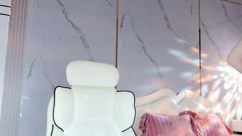 Media: A video of a modern bedroom with a white leather massage chair, a white bed with pink pillows, and a marble-patterned wall. Sunlight streams in, casting soft shadows.