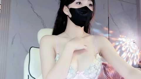 Media: Video of an East Asian woman with long black hair, wearing a black mask and a pastel lace bra, sitting in a white chair, indoors with marble walls.