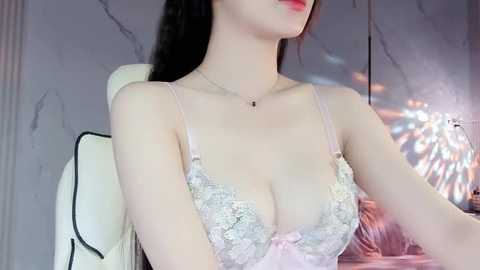 Media: Video of an Asian woman with long black hair, fair skin, and red lipstick, wearing a revealing lace bra with pink accents, seated on a white gaming chair, in a dimly lit room with marble wall and soft lighting.