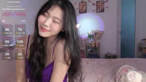 Media: A young Asian woman with long black hair and fair skin, wearing a purple dress, smiles happily in a cozy, pink-themed bedroom.