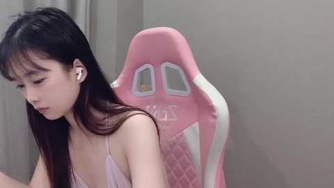 Media: A video of an East Asian woman with long black hair, wearing a pink tank top, sitting in a pink and white gaming chair, looking down thoughtfully.