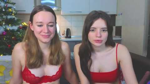 Media: Video of two young women with fair skin, one with light brown hair and red lace bra, the other with dark hair and red tank top, seated in a modern kitchen with a decorated Christmas tree.