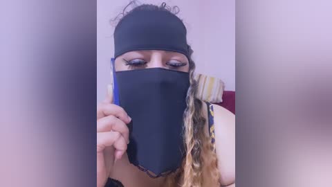 Media: Video of a woman with medium-brown skin, wearing a black face mask, holding a blue object. She has long, wavy hair with blonde highlights. The background is a blurred, neutral-colored room with a red cushion.