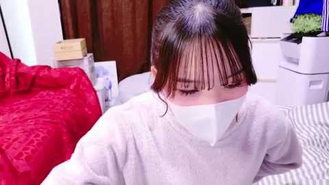 Media: A video of an Asian woman with dark hair, wearing a white face mask, a light gray sweater, and sitting on a striped bed. Background includes a red bedspread, a wooden door, and a white printer.