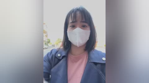 Media: Video of a young Asian woman with medium-length black hair, wearing a white surgical mask, dark blue jacket, and pink shirt, indoors with blurred background.