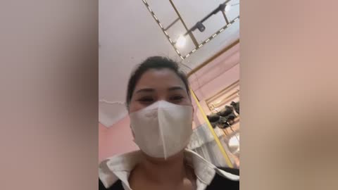 Media: Video of a woman with dark hair wearing a white surgical mask, standing in a room with pink walls and a mirror reflecting a partially visible person in the background.