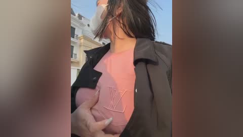 Media: Video of a person with a pink shirt, black jacket, and face mask, being held from behind by a hand, outdoors with a blurred background of buildings.