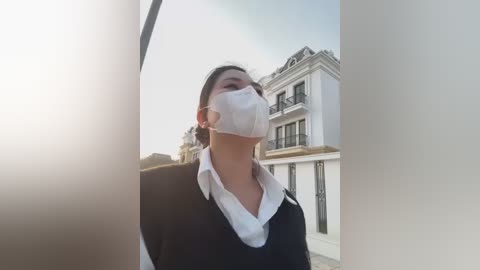 Media: Video of a woman in a black sweater and white shirt, wearing a white surgical mask, standing outdoors in front of a white mansion with a black wrought iron balcony.