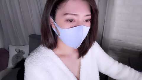 Media: Video of an East Asian woman with shoulder-length straight brown hair, wearing a light blue face mask and white fuzzy bathrobe, indoors with grey curtains and a teddy bear.