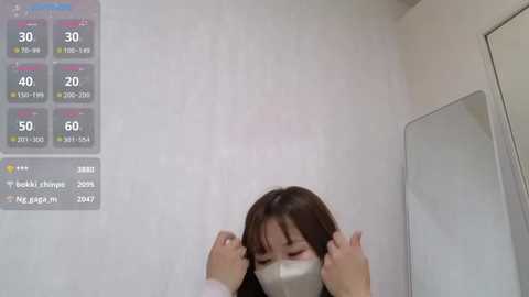 Media: A video of a woman in a white face mask adjusting her hair in a bathroom with a digital thermometer and mirror.