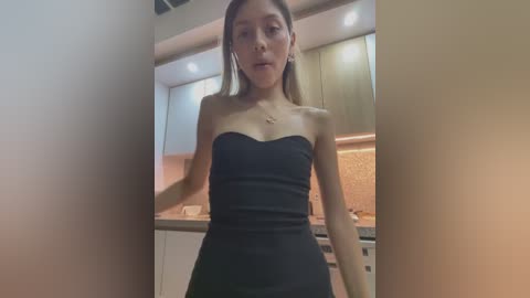 Media: Video of an Asian woman with light skin, wearing a black strapless dress, standing in a modern kitchen with beige cabinets and a backsplash. She has straight, shoulder-length hair and is looking directly at the camera.