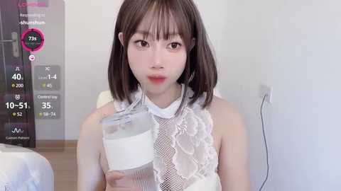 Media: Video of a young East Asian woman with straight, shoulder-length dark hair, fair skin, and wearing a white lace dress, standing indoors.