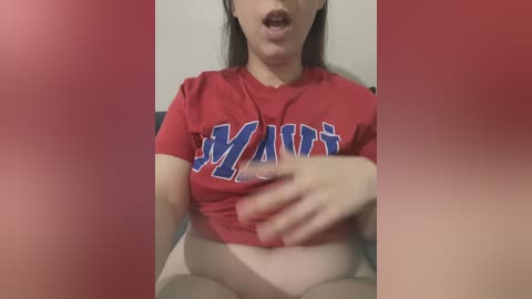 A video of a fair-skinned woman with long dark hair, wearing a red \"MVP\" T-shirt, lifted to reveal her bare stomach and crotch. The background is blurred, with red filter effects.