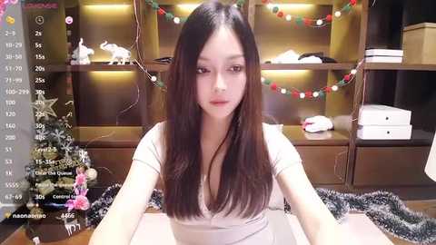 Video of an East Asian woman with straight black hair, wearing a white top, sitting at a desk with Christmas decorations and a fur blanket in the background.