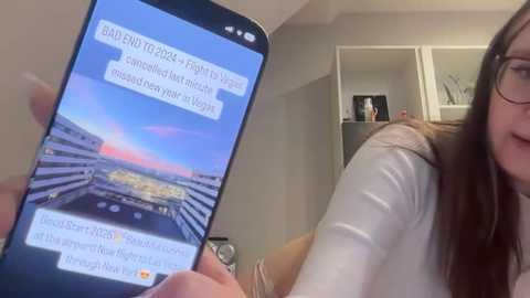 Media: Video of a woman with long brown hair, wearing glasses, holding a smartphone displaying an airplane window view, in a modern, beige-colored room.