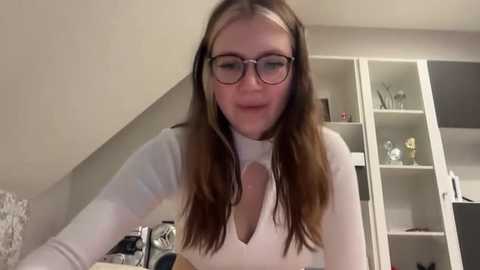 Media: Video of a fair-skinned woman with long brown hair and glasses, wearing a white top with a cut-out design, standing in a cluttered bedroom with a white wardrobe and a small table.