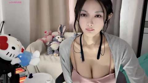 Media: Video of an Asian woman with long black hair, wearing a pink tank top and gray cardigan, sitting in a cluttered room with plush toys and a pink chair.