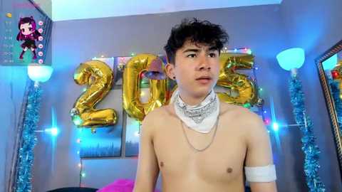 Media: Video of a shirtless, young, light-skinned man with curly black hair, wearing a white bandage on his right arm, surrounded by blue tinsel and large gold \"2015\" letters on a gray wall.