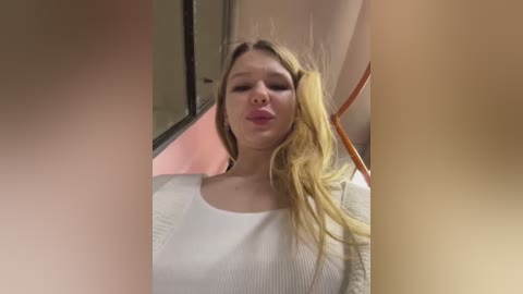 Media: Video of a blonde Caucasian woman with long hair, wearing a white sweater, making a kissy face in an elevator with beige walls and a pink background.