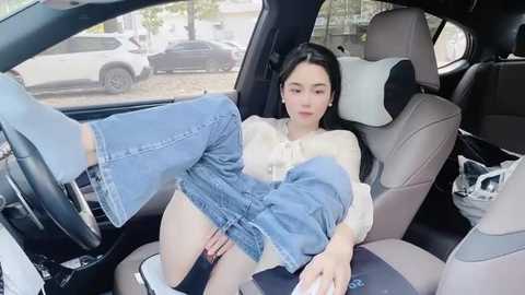 Media: Video of a young Asian woman with long black hair, wearing blue jeans and a white top, sitting in a car with the door open, legs outside, in a parking lot.