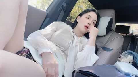 Media: Video of a young Asian woman with fair skin and long black hair, wearing a white blouse, lying in a car seat, legs spread, revealing underwear, looking tired.