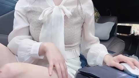 Media: Video of a fair-skinned woman with a slender build, wearing a white blouse and beige sweater, seated in a car, with a dark-haired person's bare leg visible in the foreground.