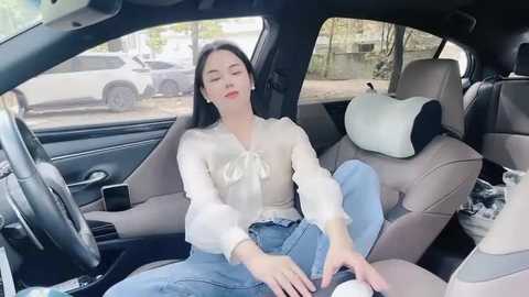 Media: Video of a young Asian woman with long black hair, wearing a white blouse and blue jeans, relaxing in a beige car seat, surrounded by parked cars outside.