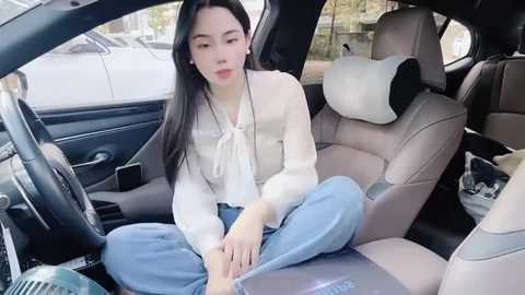 Media: Video of a young Asian woman with long black hair, fair skin, wearing a white blouse and blue jeans, sitting in a car seat with beige upholstery.