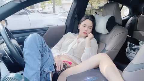 Media: Video of an Asian woman with long black hair, wearing a white blouse and blue jeans, sitting tiredly in a car seat, clutching a pink phone.