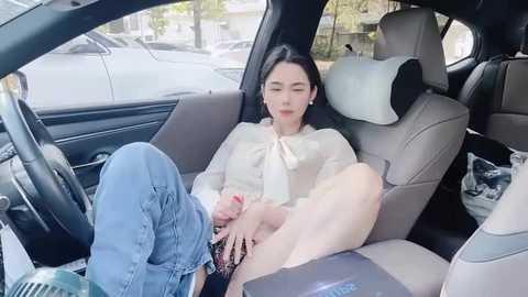 Media: Video of a young Asian woman with fair skin, wearing a white blouse and blue jeans, reclining in the backseat of a car, looking relaxed.