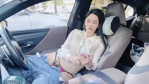 Media: Video of a young woman with long black hair, wearing a white shirt and denim shorts, asleep in the backseat of a car, surrounded by urban surroundings.