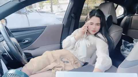 Media: Video of a young East Asian woman with long black hair, wearing a white blouse and beige pants, talking on a cellphone while sitting in a car, with a blurred street scene visible outside.