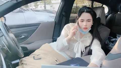 Media: Video of an Asian woman with long black hair, wearing a white shirt and beige jacket, inside a car. She's on a phone, with a beige bag on the seat beside her.