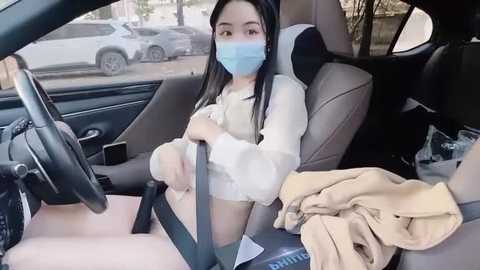 Media: Video of a young woman with long black hair, light skin, and a blue face mask, sitting in a car seat with beige clothes, holding a steering wheel.